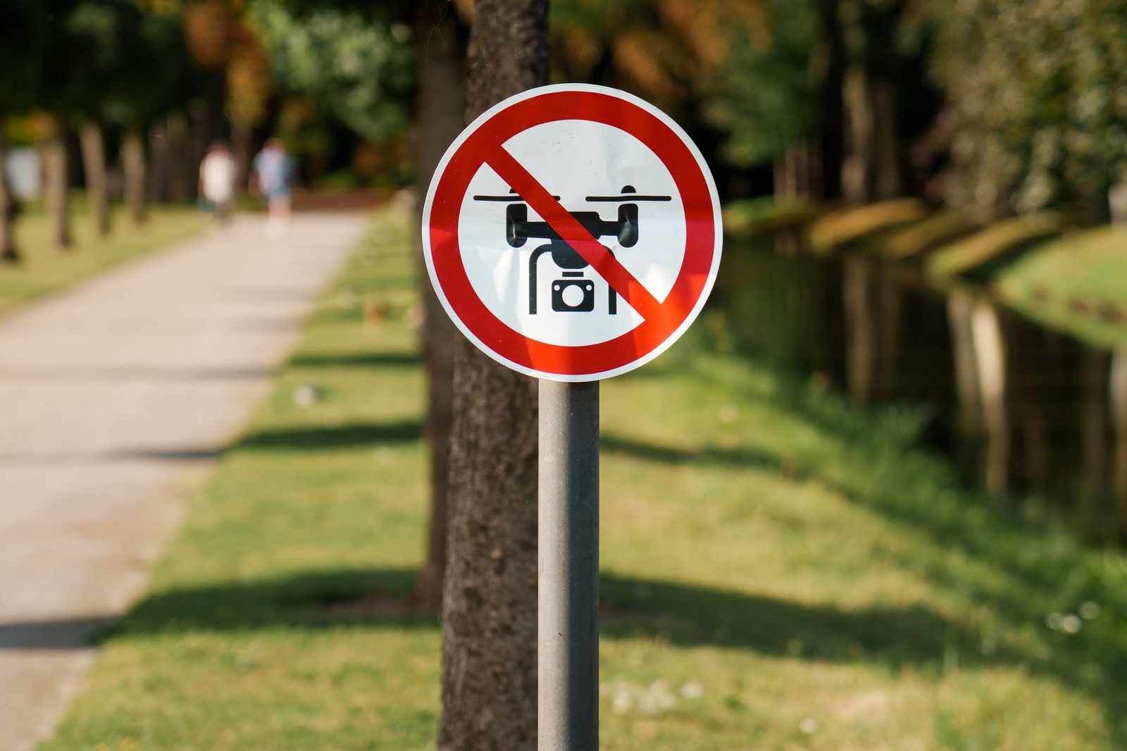 A red-bordered sign prohibiting drones, mounted on a post beside a tree-lined park path.