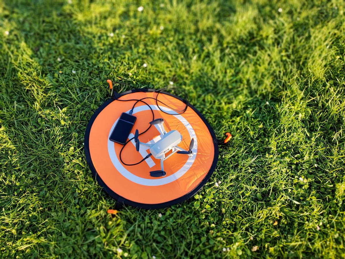 A folded drone and a phone resting on an orange landing pad in grass, seen from above.