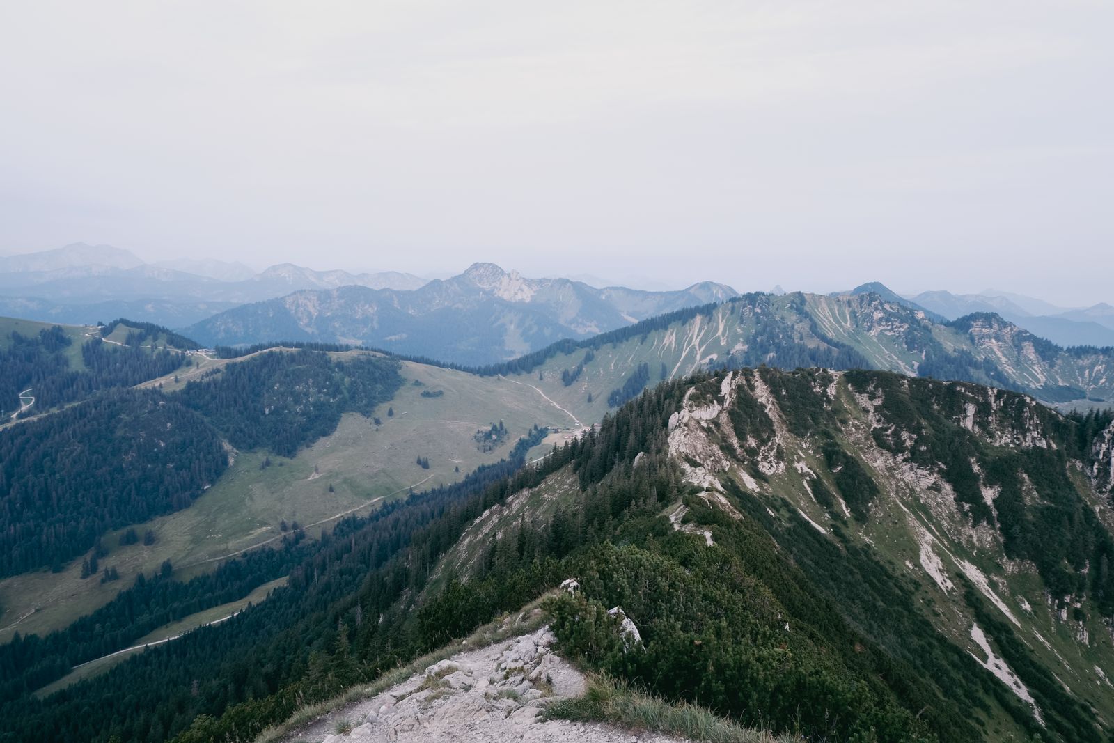 Green ridges and rocky peaks in the Bavarian Alps under a hazy summer sky.