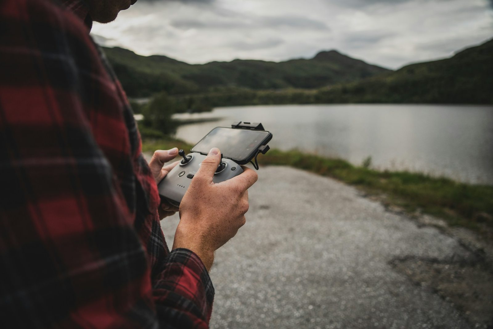 A pilot holding a drone controller with a phone clipped to it, beside a lake ringed by hills.
