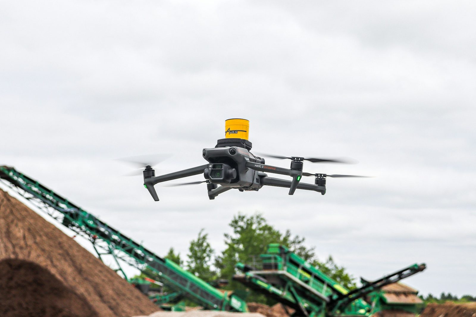 A drone carrying a yellow strobe beacon, flying above machinery on a worksite.