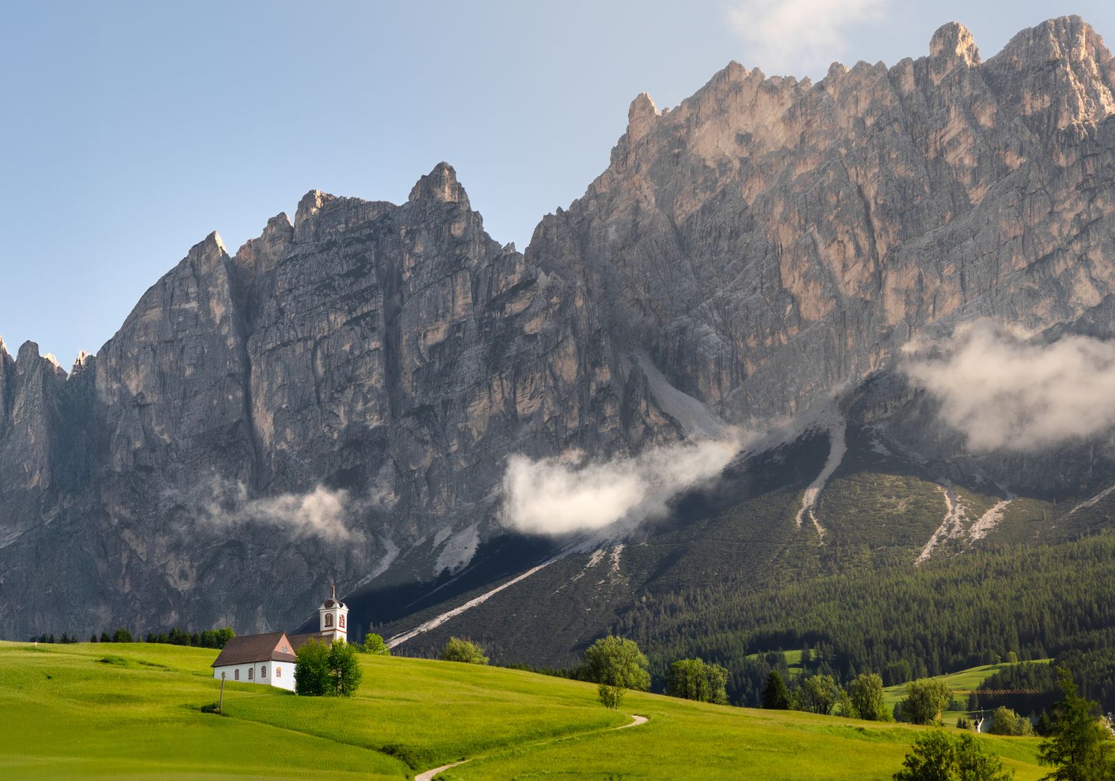 A small church and green meadow below steep rocky peaks in the Italian Dolomites.