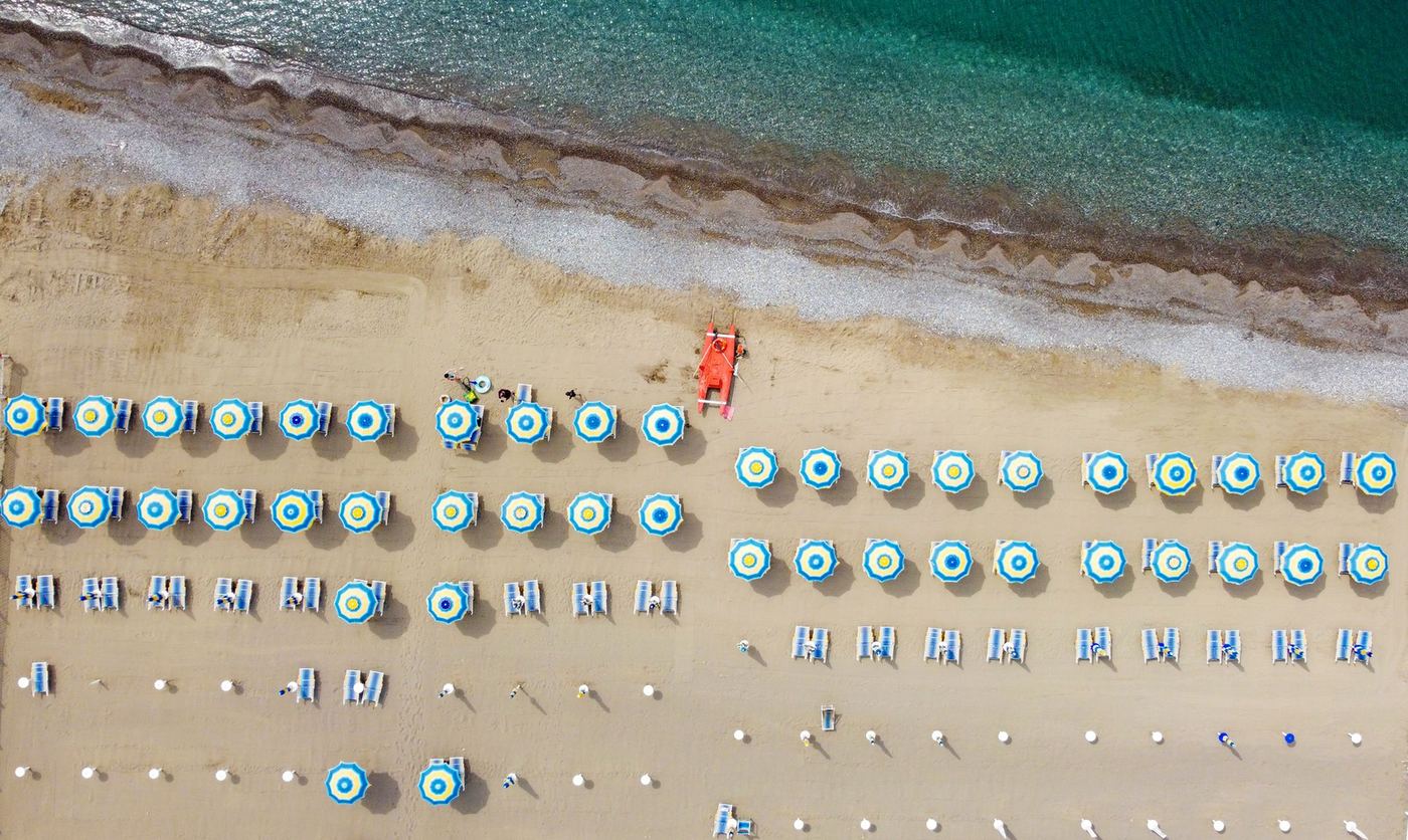 An aerial view of rows of sun umbrellas and loungers on a busy beach beside turquoise water.