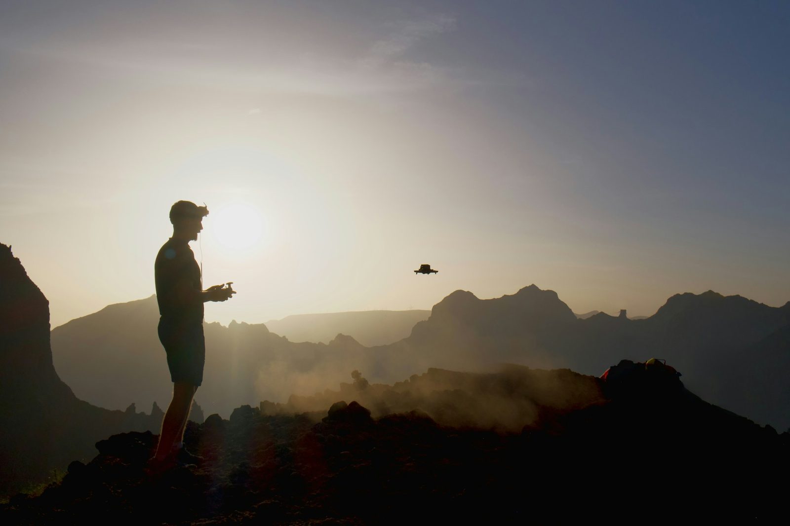 A pilot silhouetted against the setting sun on a ridge, flying a drone above a hazy mountain landscape.