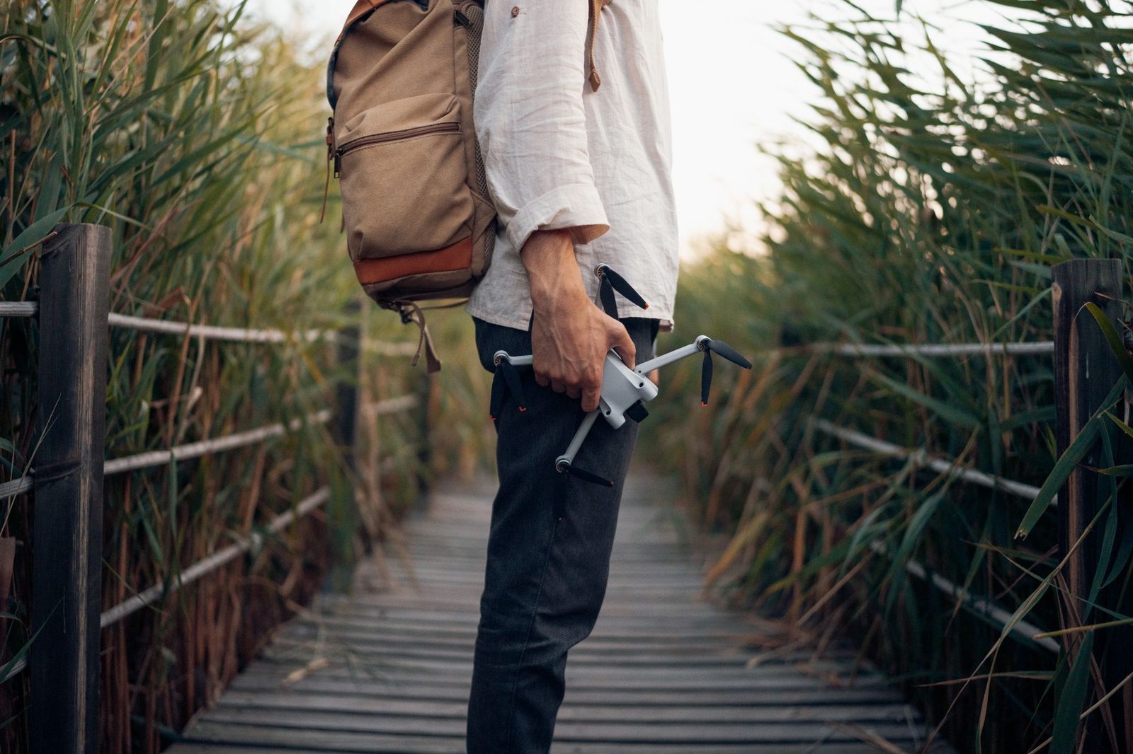 A traveller with a backpack carrying a folded drone in one hand, on a boardwalk through tall reeds.