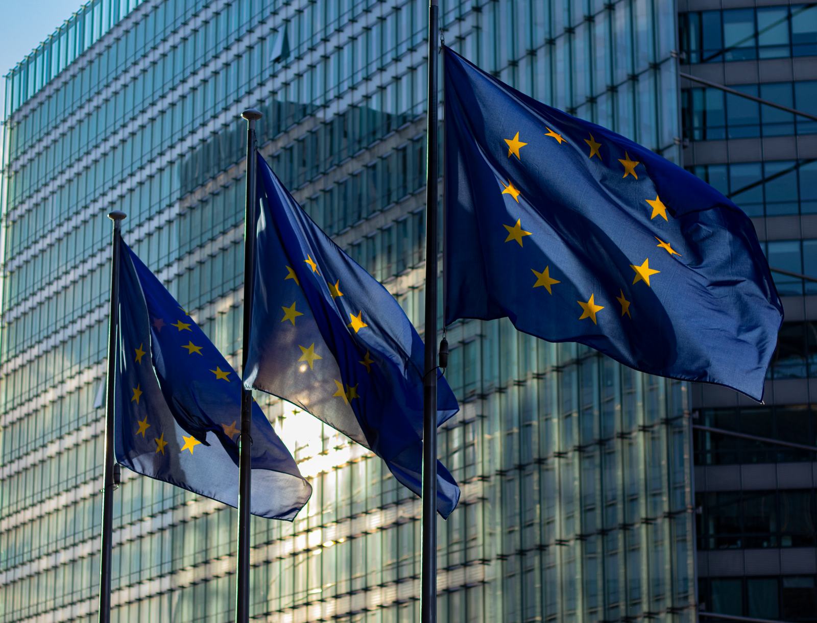 Three European Union flags flying outside a glass office building.