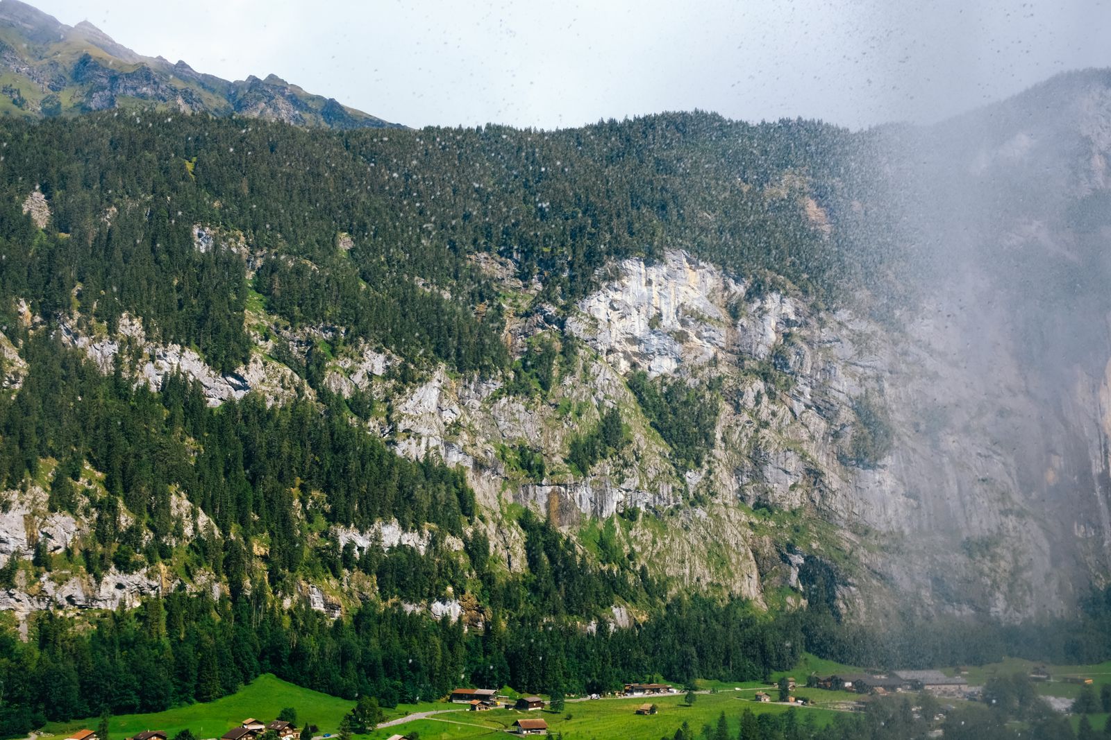Rain and low cloud crossing forested cliffs above green fields in Lauterbrunnen valley, Switzerland.