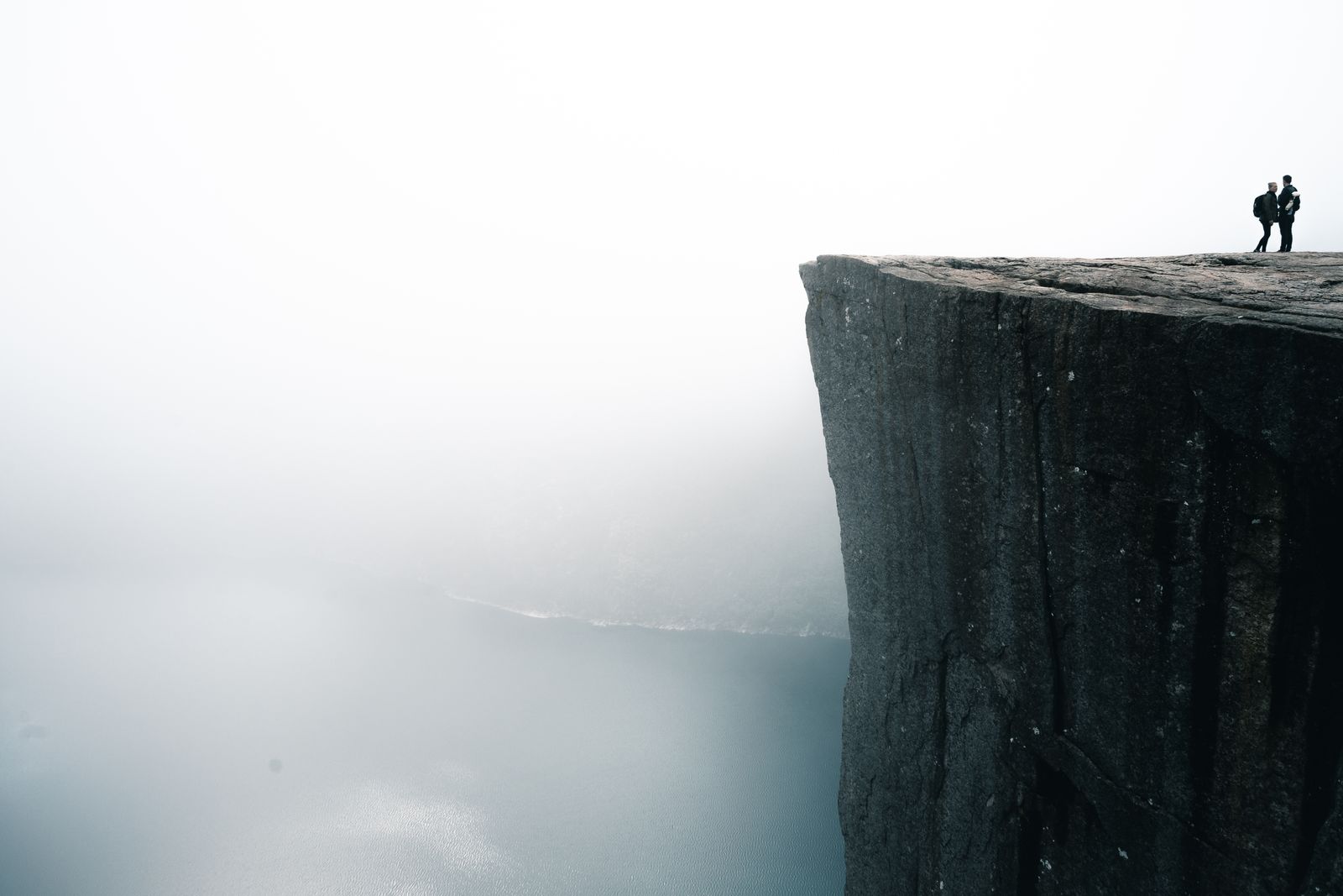 Two hikers standing on the Preikestolen cliff above a misty Lysefjord in Norway.
