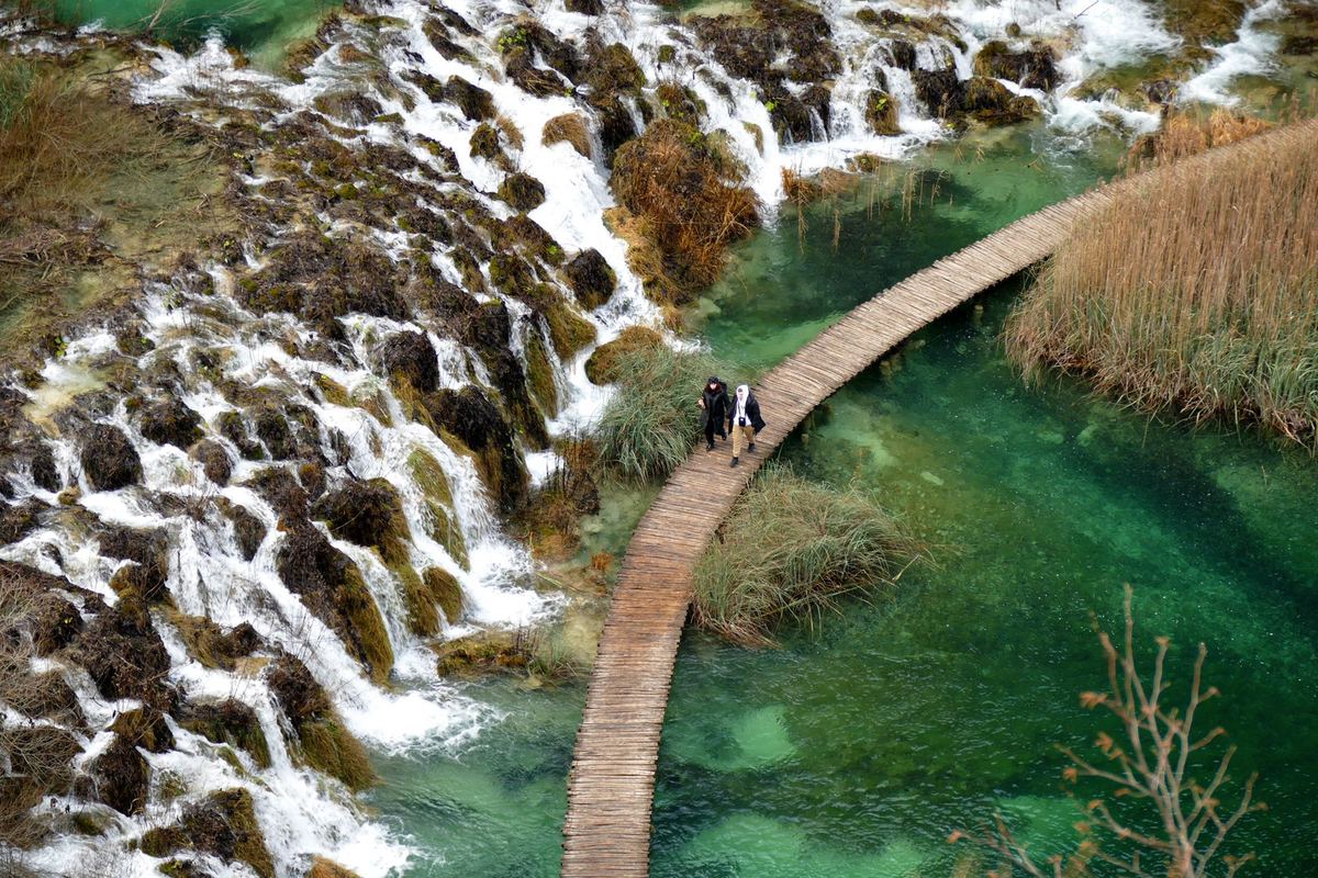 An aerial view of the terraced cascades of the Plitvice Lakes, with a wooden walkway crossing the water.