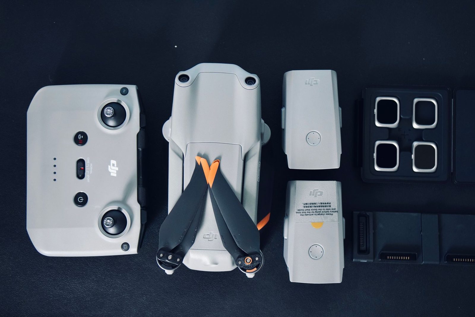 A drone controller, folded drone, spare batteries, filters and cases laid out in rows on a dark surface.