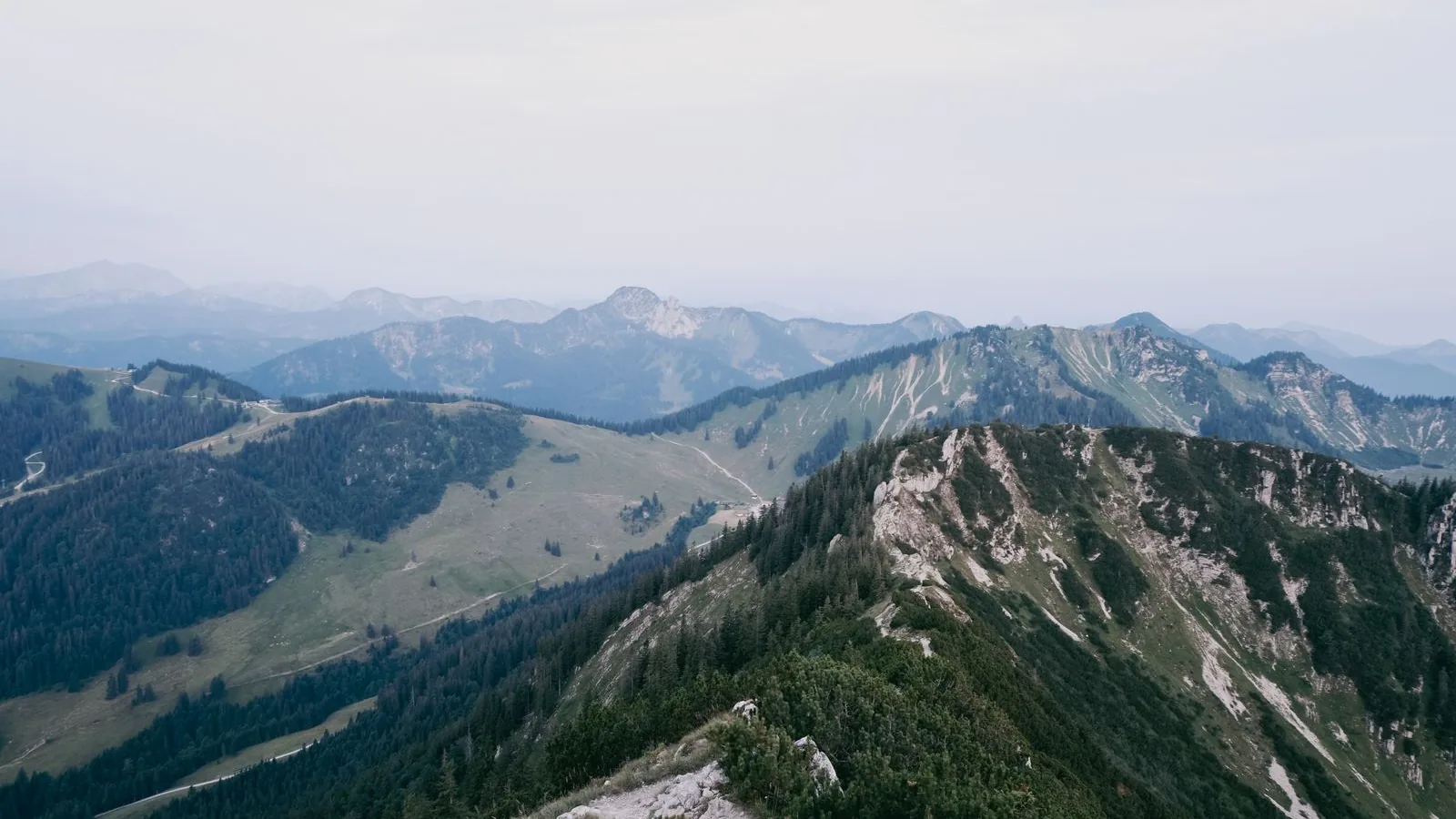 Green ridges and rocky peaks in the Bavarian Alps under a hazy summer sky.