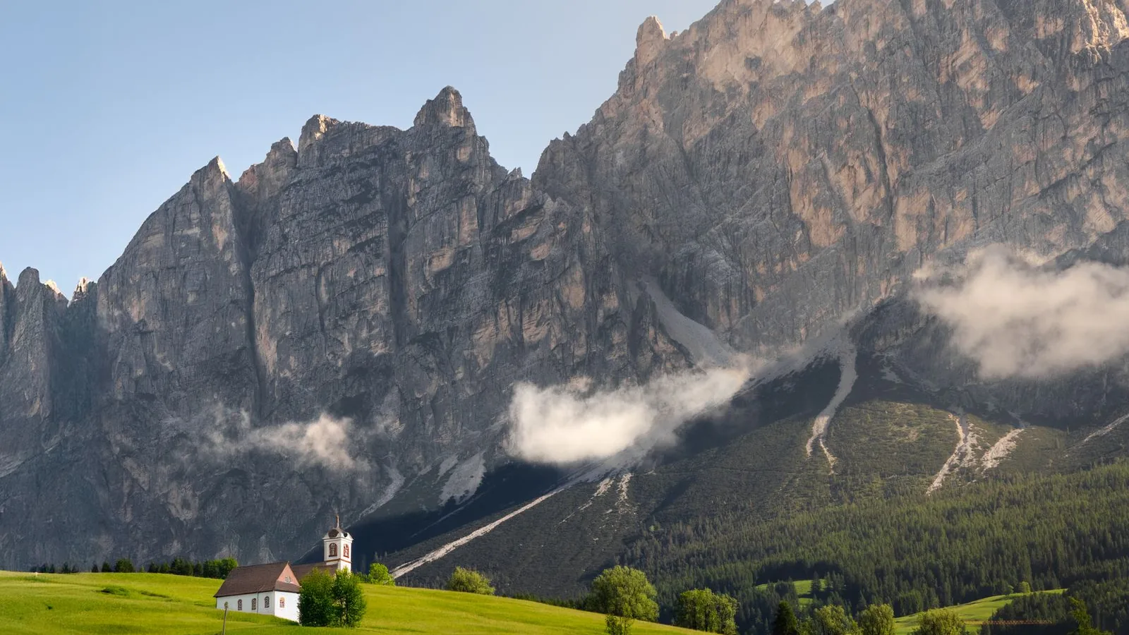 A small church and green meadow below steep rocky peaks in the Italian Dolomites.