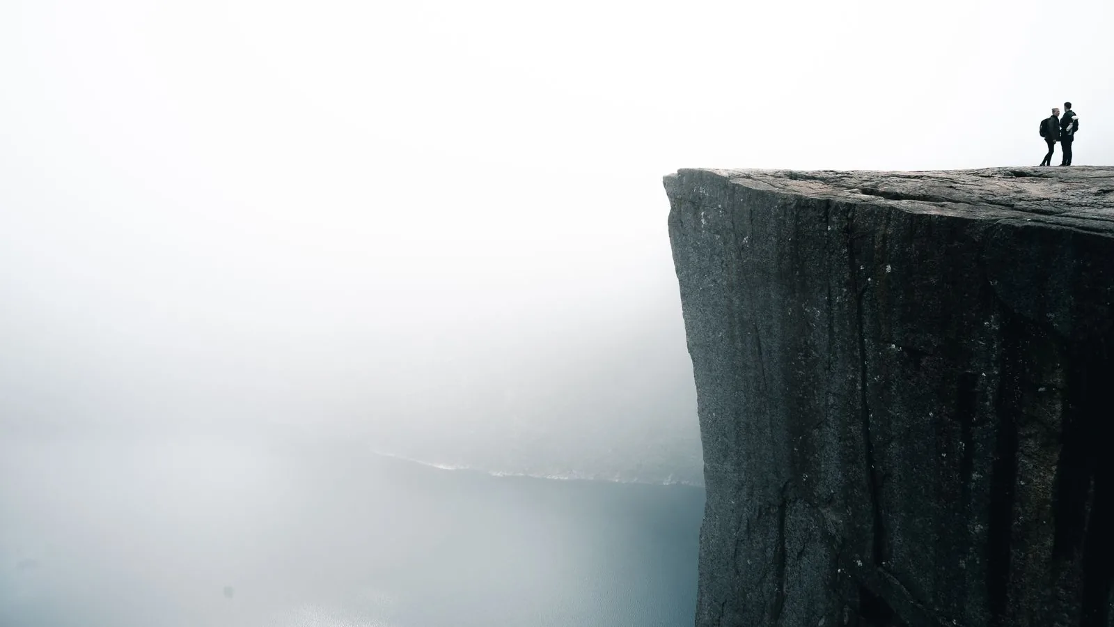 Two hikers standing on the Preikestolen cliff above a misty Lysefjord in Norway.