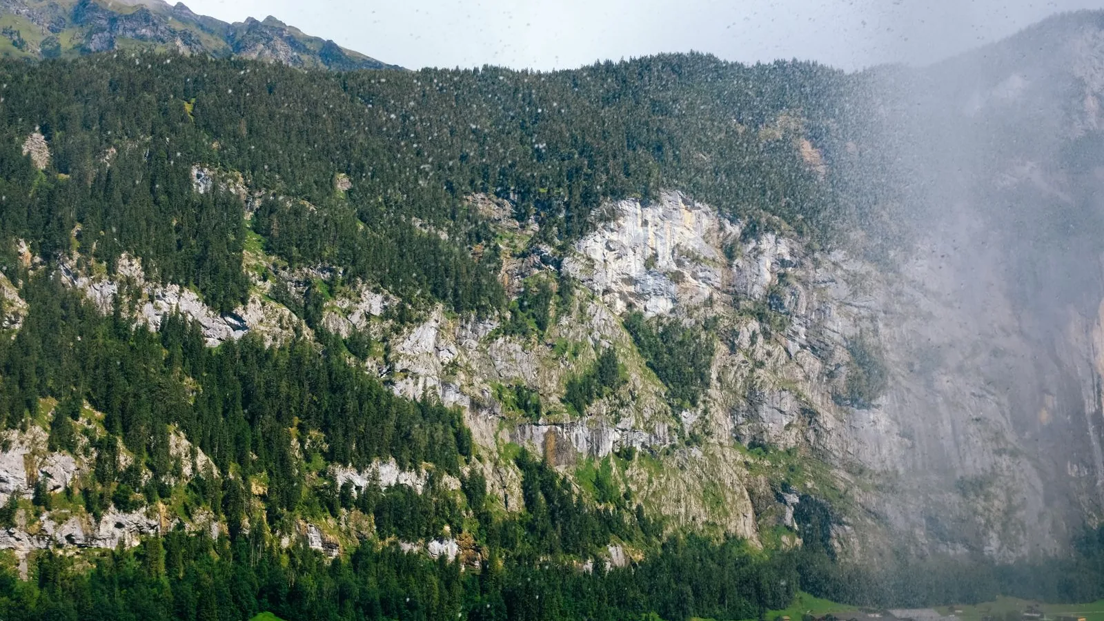 Rain and low cloud crossing forested cliffs above green fields in Lauterbrunnen valley, Switzerland.